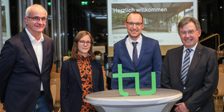 Chaudoire-Preisverleihung Eine Frau und drei Männer stehen in einer Reihe. Vor ihnen steht das TU-Dortmund-Logo auf einem Stehtisch, hinter ihnen ist eine Präsentation auf einer Leinwand zu erkennen.