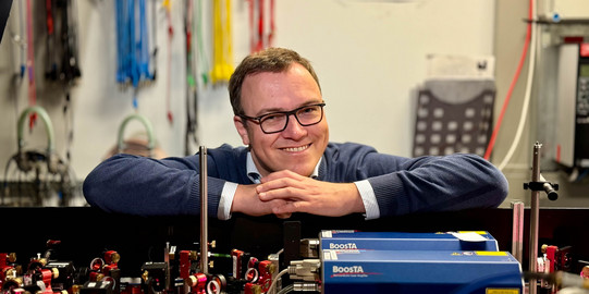 Foto: Porträt eines Manns mit Brille und blauem Pullover. Der Mann lehnt mit verschärnkten Händen auf einer Aparatur in einem Labor und lächelt in die Kamera.