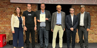 Eine Gruppe von Menschen steht nebeneinander. Ein Mann in der Mitte der Gruppe hält eine Urkunde, die von Robert Habeck (rechts im Bild) verliehen wurde.