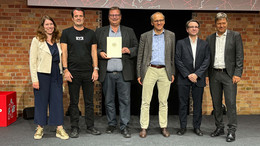 Eine Gruppe von Menschen steht nebeneinander. Ein Mann in der Mitte der Gruppe hält eine Urkunde, die von Robert Habeck (rechts im Bild) verliehen wurde.