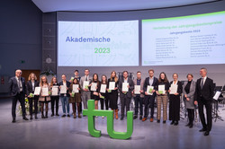 Eine Gruppe von Menschen mit Urkunden hinter dem TU-Logo bei der akademischen Jahresfeier der TU Dortmund.