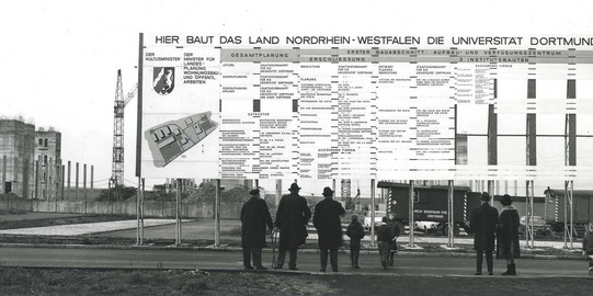 Historisches Schwarz-Weiß-Foto vor der Baustelle der TU Dortmund. Mehrere Personen betrachten ein Schild mit Informationen zum Bau der Universität.