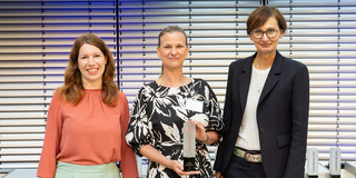 Three women pose side by side for a photo. The woman in the middle is holding an award in her hand.