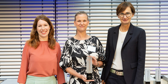 Unipreneurs Flatten Drei Frauen posieren nebeneinander für ein Foto. Die Frau in der Mitte hält einen Preis in der Hand.