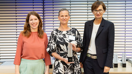 Drei Frauen posieren nebeneinander für ein Foto. Die Frau in der Mitte hält einen Preis in der Hand.