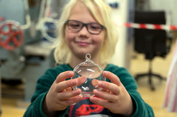 Das Bild zeigt ein Kind mit blonden Haaren und schwarzer Brille, welches mit ausgestreckten Armen eine Glaskugel in den Händen hält und lächelt diese an.