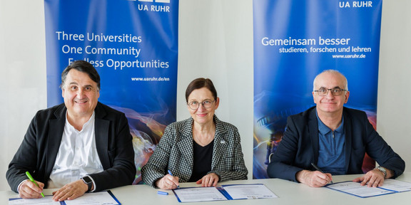 Photo: Three people sign an agreement in front of roll-up banners of the University Alliance Ruhr.