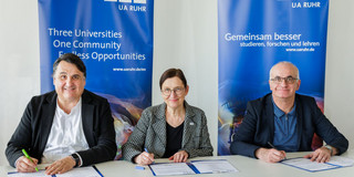 Foto: Drei Personen unterzeichnen eine Vereinbarung vor Roll‑up‑Bannern der Universitätsallianz Ruhr.