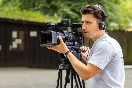 Weg in den Journalismus Ein Mann hält eine Kamera auf einem Stativ in den Hängen und trägt ein Headset.