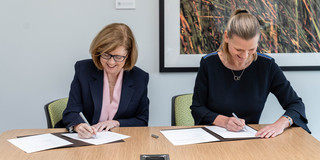 Two women in formal dress sit at a table and sign documents in brown folders.