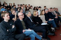 Ein Publikum von Studierenden und Dozent*innen sitzt in einem Saal und hört zu.