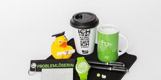 Verschiedene Merchandise-Artikel der TU Dortmund, unter anderem eine Tasse, eine Uhr und ein Schlüsselanhänger, liegen auf einem Tisch