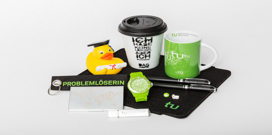 Verschiedene Merchandise-Artikel der TU Dortmund, unter anderem eine Tasse, eine Uhr und ein Schlüsselanhänger, liegen auf einem Tisch