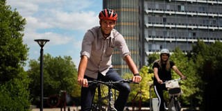 Stadtfahrrad fahren Ein Mann mit einem roten Helm sitzt auf einem Fahrrad und hält an, neben ihm fährt eine Frau mit weißem Helm auf einem Fahrrad.
