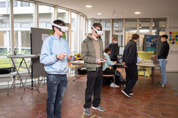 Zwei Jungen stehen im Audimax-Foyer und haben VR-Brillen auf. Hinter ihnen ein Stand mit Tischen und Aufstellern, an dem Studierende und Besucher*innen stehen.