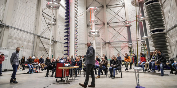 In einer Experimentierhalle stehen große Türme aus Metallapparaten. Darunter sitzt ein großes Publikum. Im Vordergrund stehen zwei Männer mit einem Beamer und halten einen Vortrag.