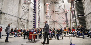 In einer Experimentierhalle stehen große Türme aus Metallapparaten. Darunter sitzt ein großes Publikum. Im Vordergrund stehen zwei Männer mit einem Beamer und halten einen Vortrag.