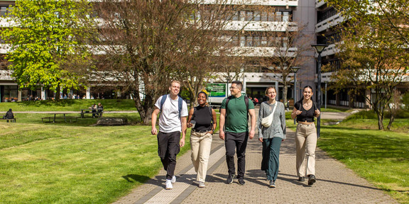 Five people walk in a row across the summer campus, with the building at Emil-Figge-Straße 50 in the background