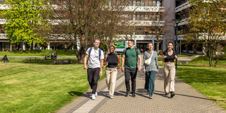 Five people walk in a row across the summer campus, with the building at Emil-Figge-Straße 50 in the background