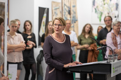 Rundgang Kunst Prof. Wiebke Möhring Prof. Wiebke Möhring steht an einem Rednerpult. Sie wendet sich der Studierenden links im Bild zu. Im Hintergrund stehen Menschen.