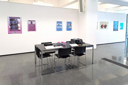 Im Zentrum des Bildes ist ein schwarzer rechteckiger schwarzer Tisch mit fünf schwarzen Stühlen zu sehen, die auf einem schwarz-grau glänzenden Boden stehen. Im Hintergrund ist eine lange weiße Ausstellungswand mit verschiedenen Leinwänden zu sehen.