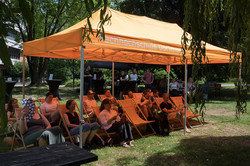 Photo: Several people sit on deck chairs under a pavilion at an outdoor event.