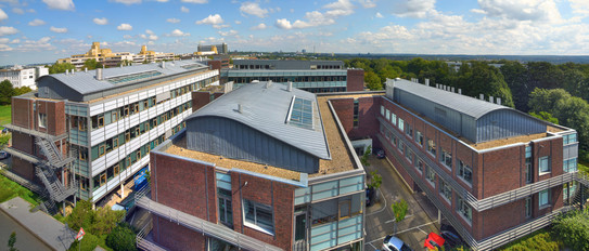 Foto: Ein Panoramabild der Gebäude des MPI. Es sind drei Backsteingebäude mit grauen Metalldächern. Im Hintergrund sind der bewölkte Himmel und der Mathetower der TU Dortmund zu sehen.