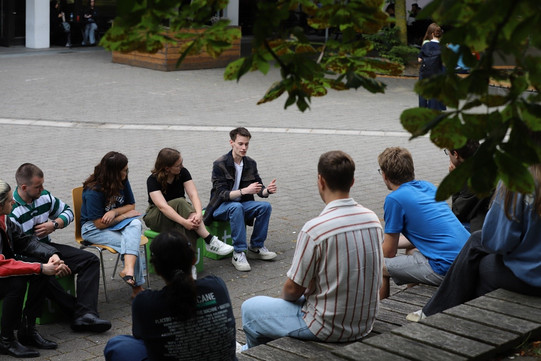Eine Gruppe von Studierenden sitzt im Freien in einem Kreis zusammen und führt ein Gespräch. Einige sitzen auf Stühlen, andere auf einer Holzbank. Im Hintergrund sind weitere Personen und Gebäude auf dem Campus zu sehen, umgeben von Grünpflanzen.