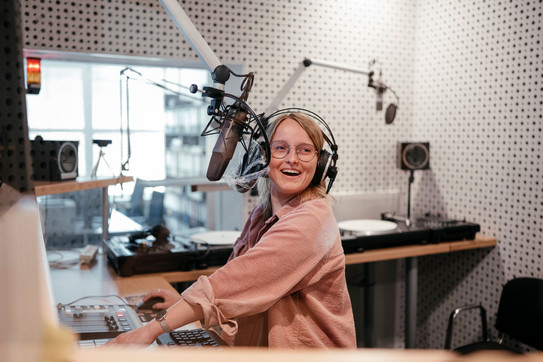 Photo: A student wearing headphones sits in front of a microphone in the campus radio studio and laughs at the camera.