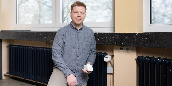 Foto: Ein blonder kurzhaariger Mann steht vor einem Heizkörper und hält ein digitales Heizkörperthermostat in der Hand.