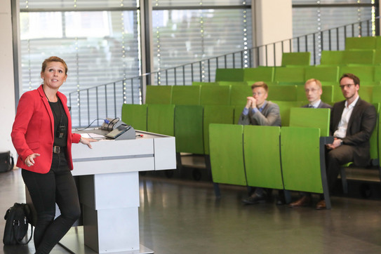 Thyssenkrupp Manager-Cup Eine Frau mit rotem Blazer steht im Hörsaal und hält einen Vortrag. Im Hintergrund sitzen drei Männer in Anzügen.