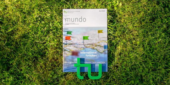 mundo_international forschen Die Ausgabe 34/2023 der Forschungszeitschrift mundo liegt im Gras, am unteren Rand liegt ein kleines, grünes TU-Logo auf der Zeitschrift.