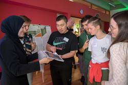 A woman in a black hijab smiles and holds a folder of photos through which a young man with short black hair and a black T-shirt is leafing through. Next to him are other young people who are also looking at the pictures.