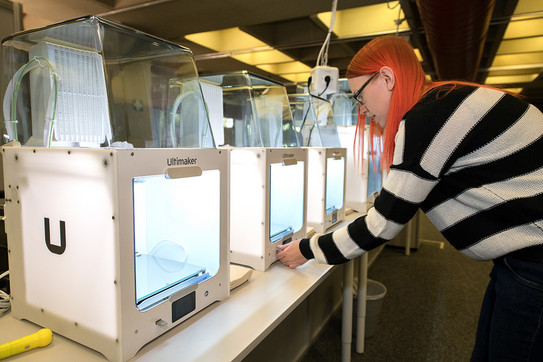 Eine Frau steht vor eine Reihe von 3D-Druckern, die aufgereiht auf einem Tisch stehen.