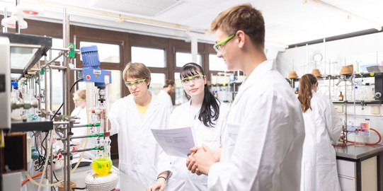 Drei Personen in Laborkitteln stehen in einem chemischen Labor und besprechen etwas.