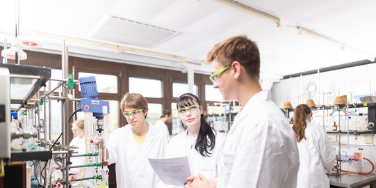 Three people in lab coats are standing in a chemical lab discussing something.
