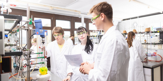 Three people in lab coats are standing in a chemical lab discussing something.