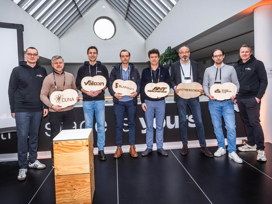 Das Bild zeigt die sechs ersten Start-ups des innoclub. Sie stehen in einer Reihe und lächeln in die Kamera. Zu sehen ist außerdem der schwarze Boden, eine Art Pult sowie ein spitz zulaufendes Dach.