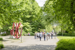 Mai Mehrere Studierende gehen über einen Weg auf dem Campus Nord im Sommer und grüne Bäume stehen rechts und links.