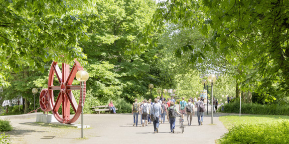 Campus North Several students walk across a path on the North Campus in the summer and green trees stand to the right and left.