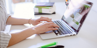 There is a laptop on a desk. You can see hands working on the laptop.