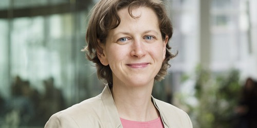 Eine Frau mit kurzem, hellbraunen Haar steht in einem modernen Gebäude mit Glasfront und Pflanzen im Hintergrund. Sie trägt ein beigefarbenes Sakko über einer rosafarbenen Bluse. Die Frau heißt Katharina Schnetgöke. 