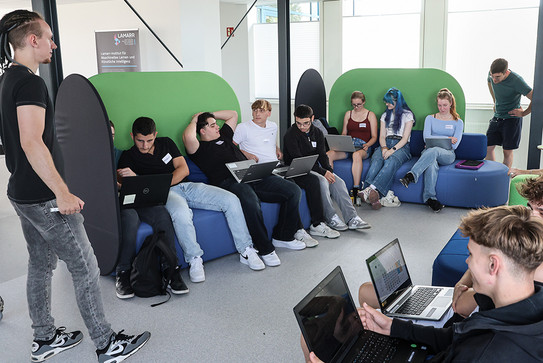 Jugendliche sitzen verteilt auf verschiedenen Sofas in einem Raum verteilt und arbeiten an Laptops.