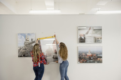 Zwei junge Frauen hängen in einem Ausstellungsraum ein Bild an eine weiße Wand, an der bereits andere Bilder hängen.