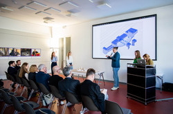 The picture was taken from the side of the room. It shows the first two rows of chairs, one of which is empty and the other is occupied by the audience. A total of five young adults are standing in front of the audience. Also visible is a drawn model in blue on a canvas and a wooden model placed on a table.