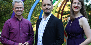 Three people stand in front of the sculpture "Spectral Rings" on the TU Dortmund campus