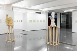 Zwei Skulpturen stehen auf Holzpodesten, im Hintergrund hängen Bilder/Kunstwerke an der Wand.