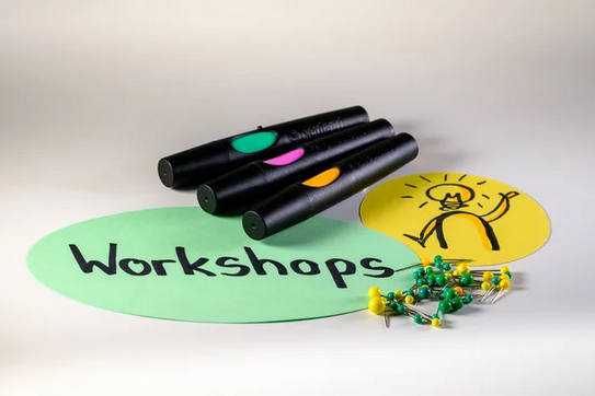 Drei schwarze Marker mit farbigen Spitzen liegen auf grüner Papierschablone mit dem Wort 'Workshops' und gelbem Kreis mit gezeichnetem Lichtsymbol, daneben grüne und gelbe Reißzwecken
