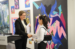 Foto: Zwei Frauen stehen vor verschiedenen Kunstwerken. Die Frau links gibt der Frau rechts die Hand und überreicht ihr eine Urkunde.