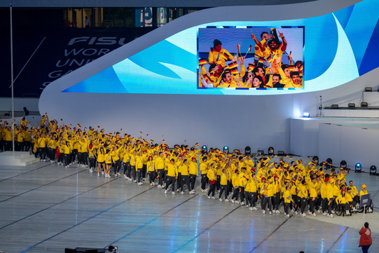 Studierende in schwarz-gelben Sportanzügen laufen bei der Eröffnungsfeier ein und halten Deutschlandfahnen in den Händen.
